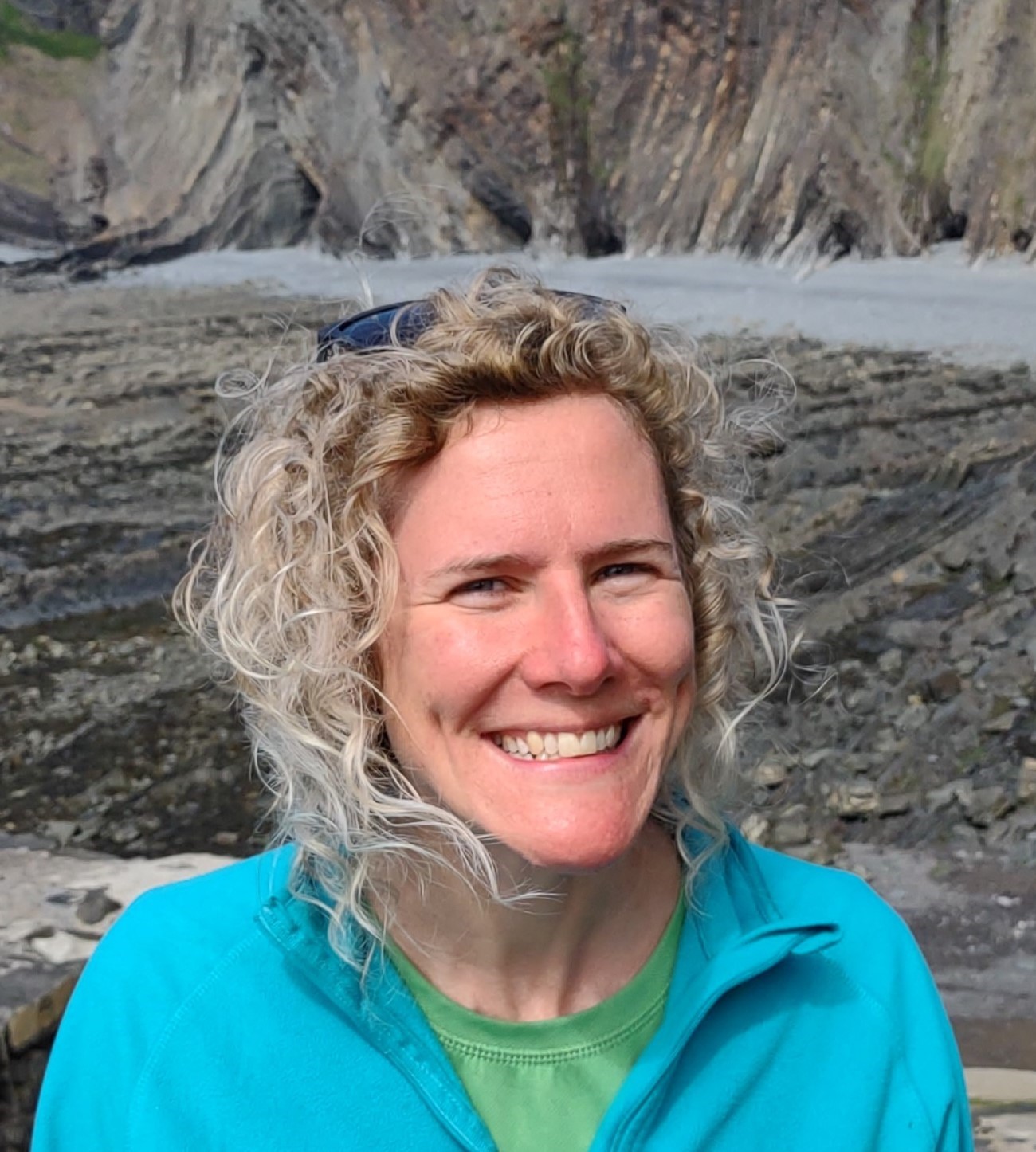 Headshot of Dr Catherine Mottram who has light coloured, shoulder length curly hair and is wearing a green t-shirt with light blue jumper over the top.
