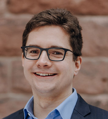 Headshot of Dr Johannes Miocic who has short hair and is wearing glasses and a blue collared shirt and jumper.