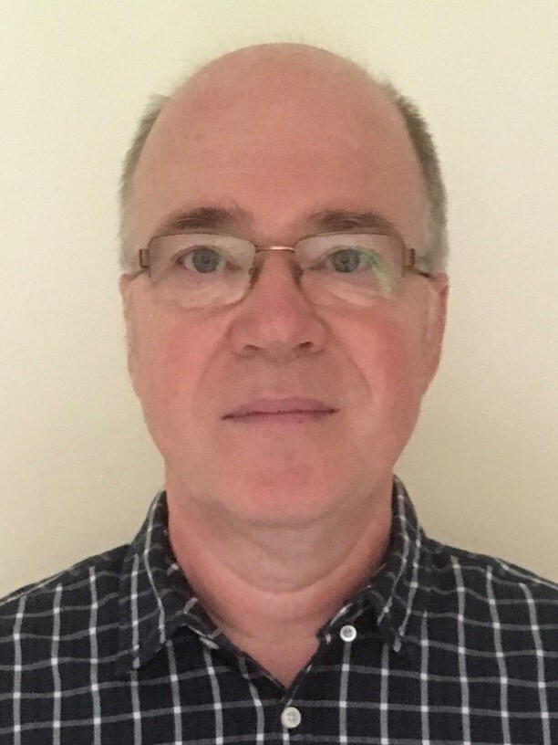 Headshot of Tom Mckie who is bald and is wearing glasses and a dark collared shirt.