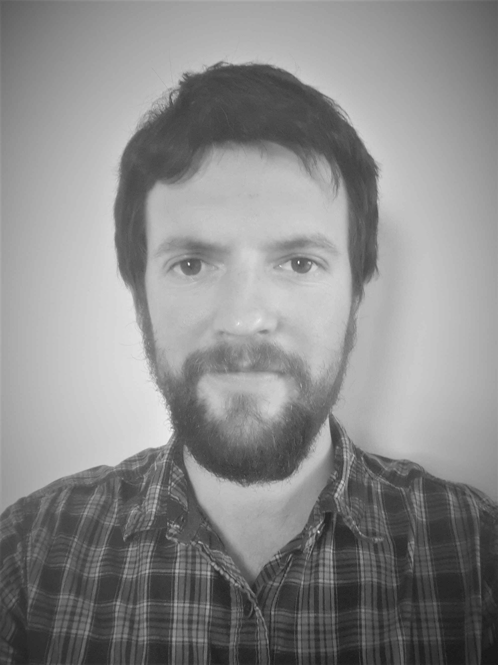 Headshot of John Millet who has dark hair and a dark beard. and is wearing a dark shirt.